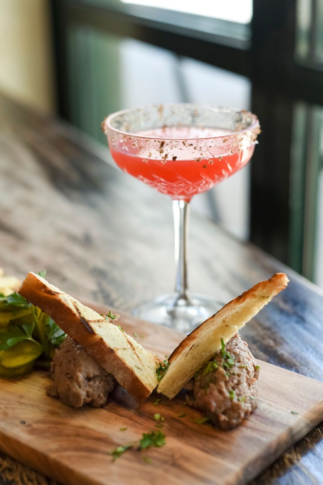 Cocktail in a coupe glass beside a plated appetizer on a wooden board by a window.