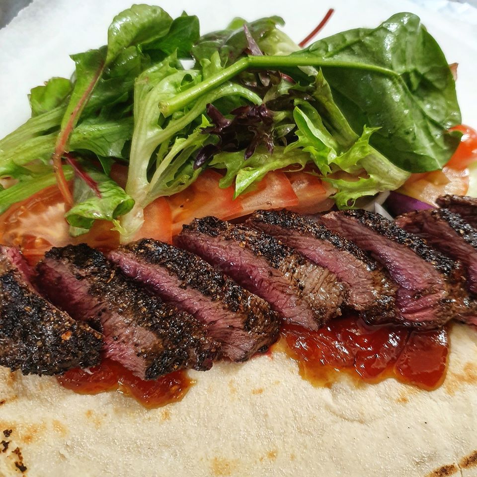 Sliced grilled steak on a wrap with red sauce, tomatoes, and mixed greens