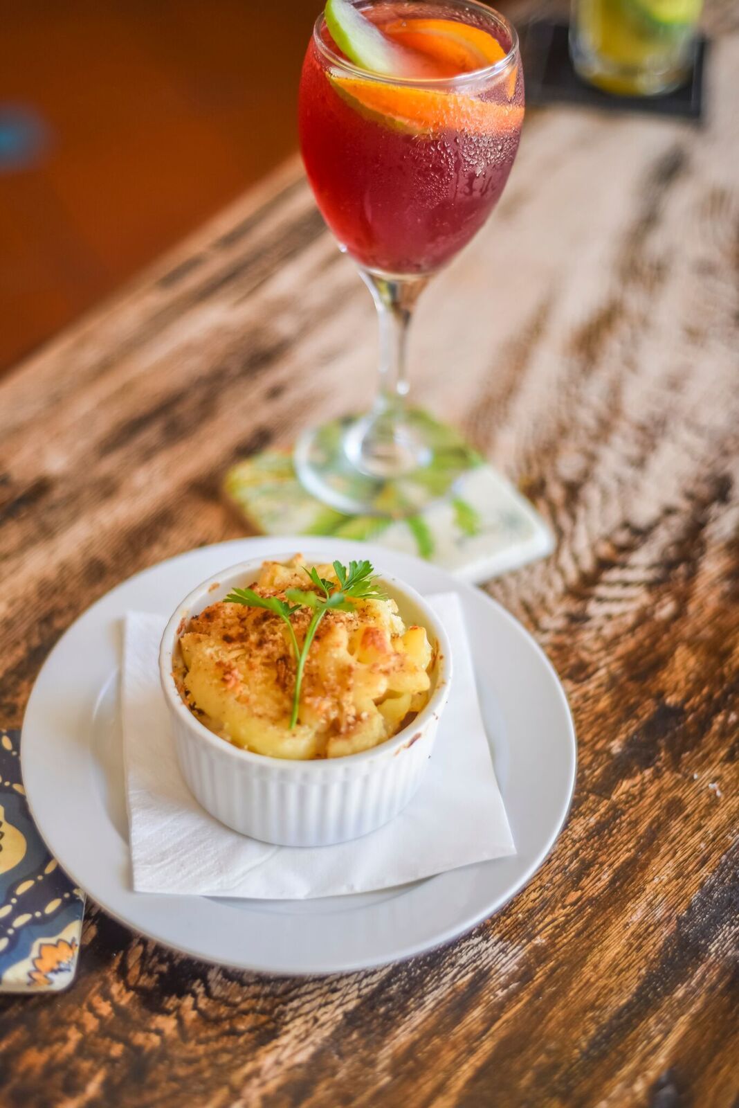 Red cocktail with orange slice beside a baked appetizer in a white ramekin on a wooden table