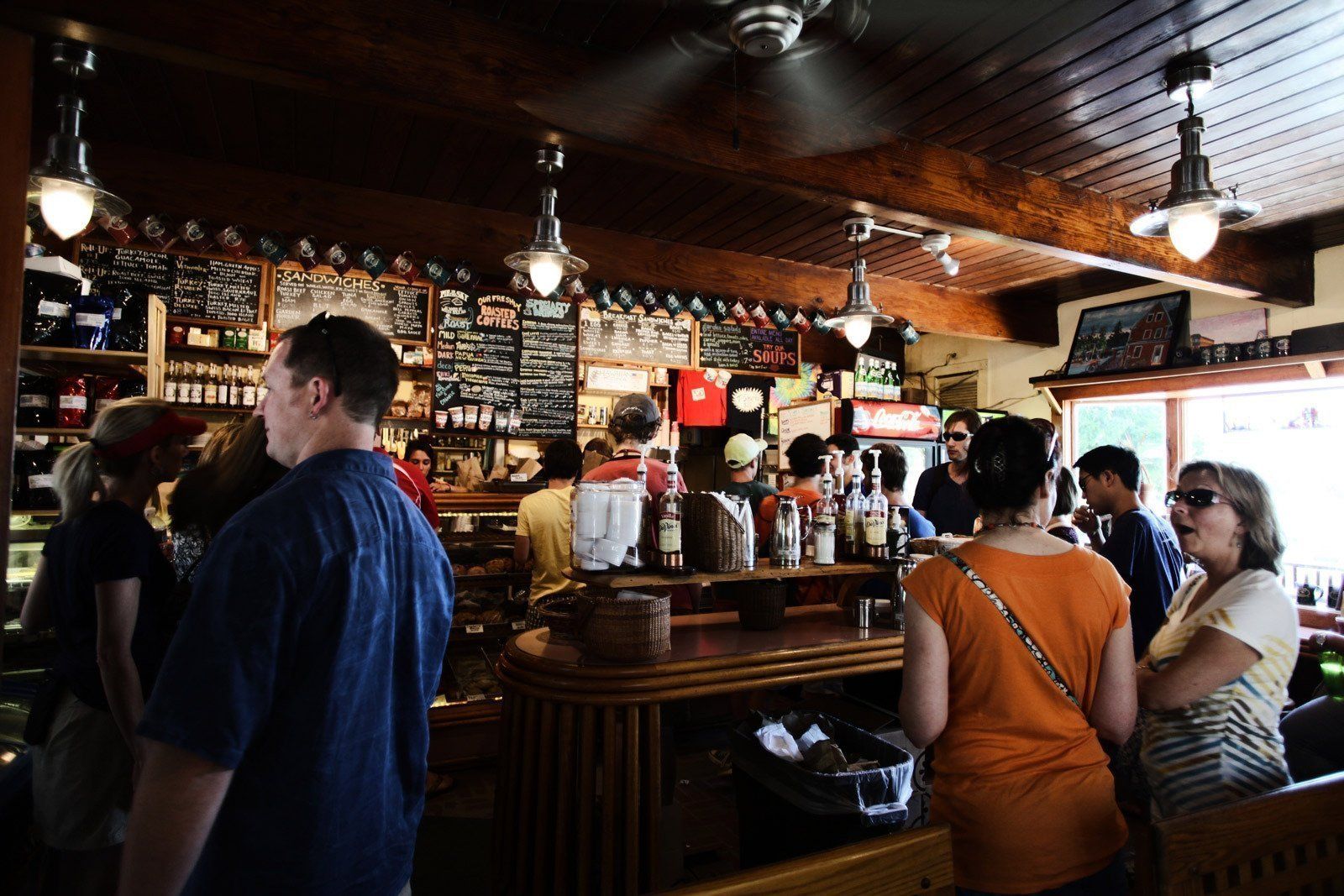 Crowded bar interior with people standing at the counter beneath hanging lights and chalkboard menus