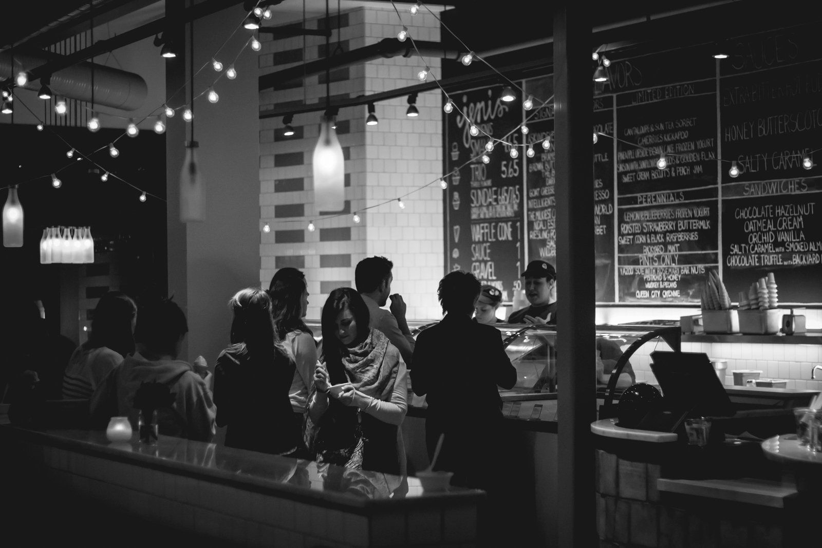 Crowded bar or café at night, with people seated and standing beneath hanging lights and chalkboard menus.