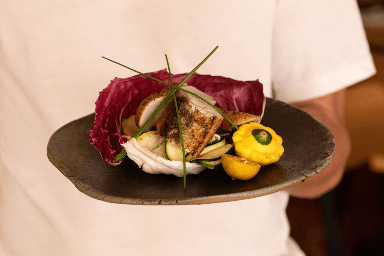 Person holding a black plate with a gourmet fish dish, yellow garnish, and dark red leaves