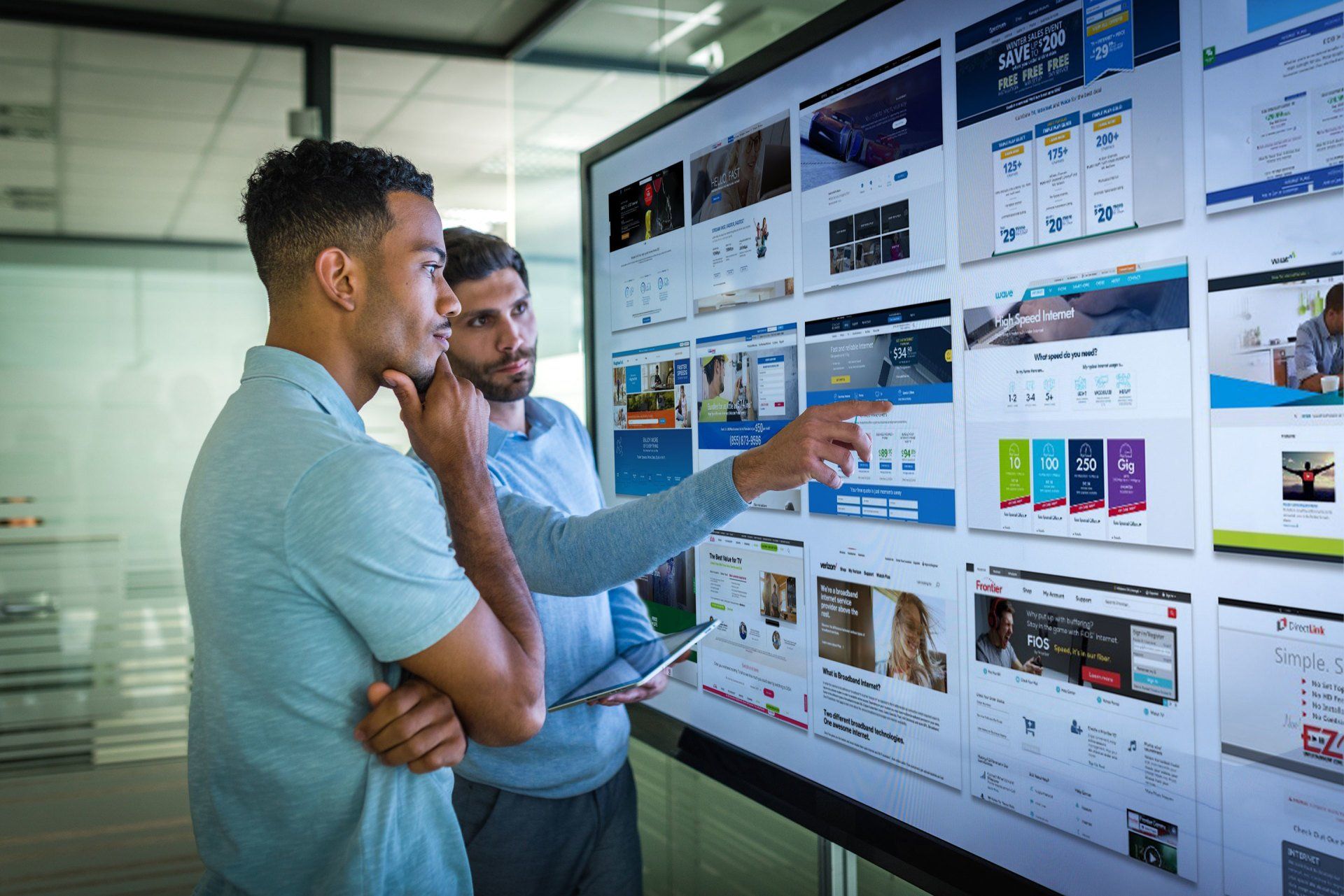 Two men are looking at a large screen with a lot of pictures on it.