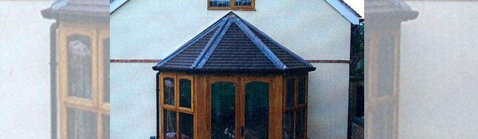 A light-colored house featuring a wooden conservatory extension with a dark tiled, multi-faceted roof.