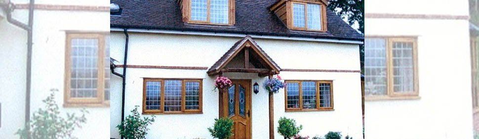 A white two-story cottage with a brown wooden door, gabled porch, small flower baskets, and wooden-framed windows.