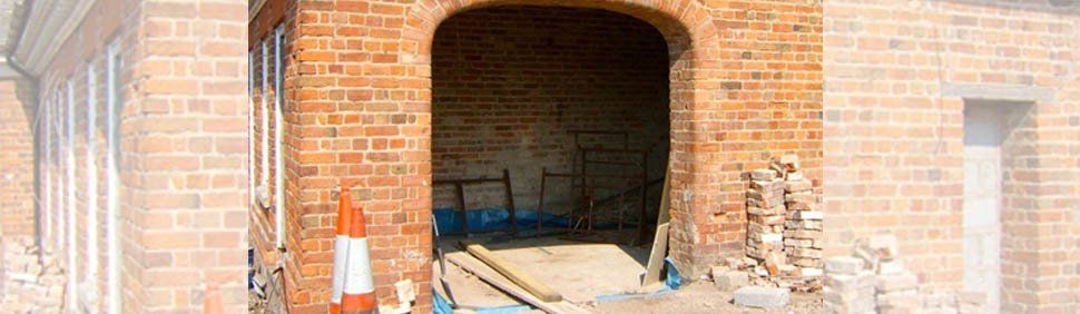 A brick building under renovation with a large arched doorway, construction equipment, and a pile of bricks.