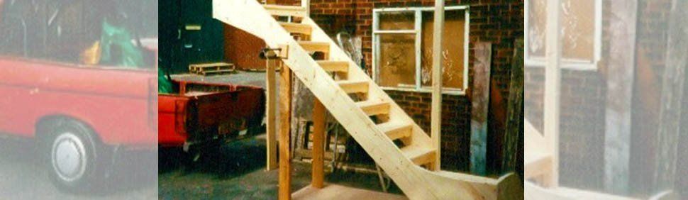 A partially assembled wooden staircase stands in a workshop, next to a red vehicle and a brick wall.