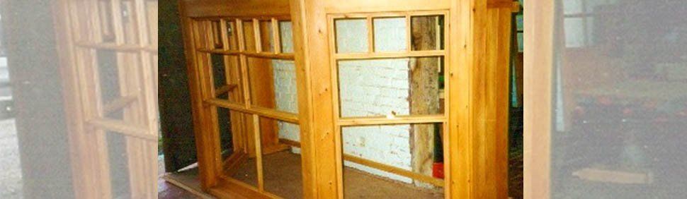 A natural-finish wooden window frame under construction, featuring multiple panes and vertical slats in a workshop setting.