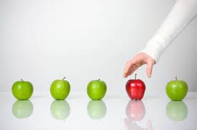 A hand is picking up a red apple from a row of green apples.