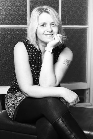 Woman with blonde hair, smiling, seated with hand on chin, wearing a patterned top, leggings, and boots.