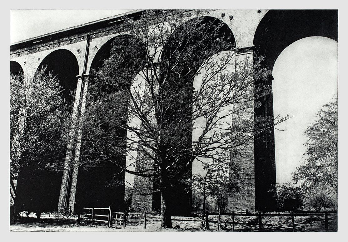 Black and white photo of a tall stone viaduct with arches. Trees and a fence stand below.