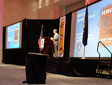 “Speaker presenting at a conference on a stage with large screens displaying event information.”