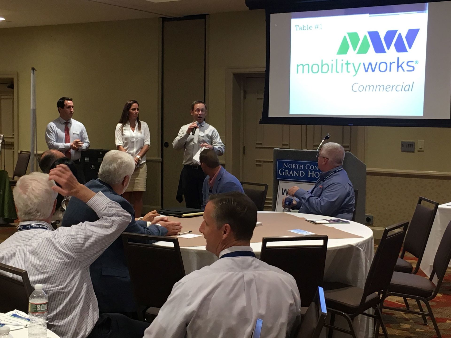 “Presenter speaking to a group at a conference while a slide about MobilityWorks Commercial is displayed on a screen.”
