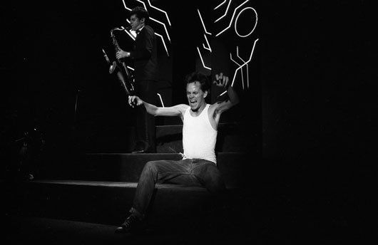 A man in a white tank top is sitting on a stage with his arms outstretched.