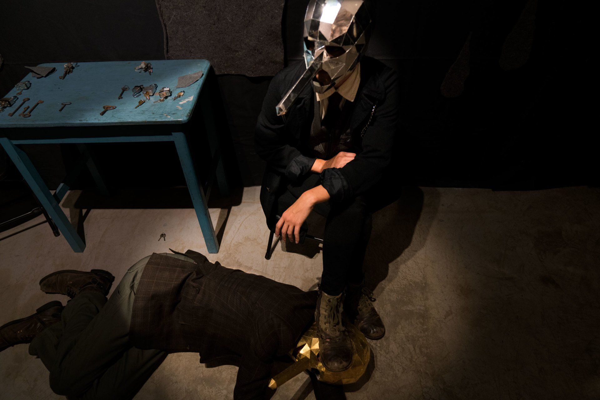 A man in a mask is sitting on a chair next to a man laying on the floor.