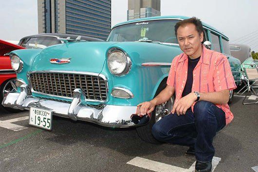A man in a pink shirt is kneeling in front of a blue car.
