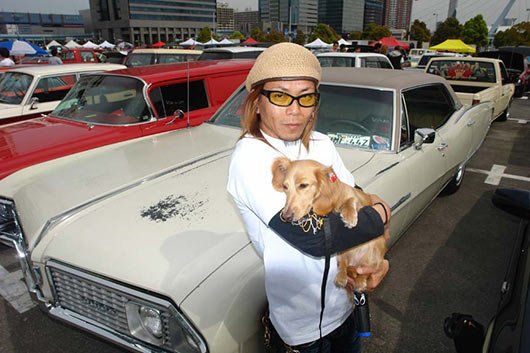 A woman is holding a small dog in front of a row of cars.