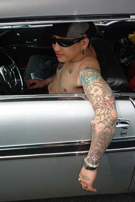 A shirtless man with tattoos on his arms is sitting in a car.