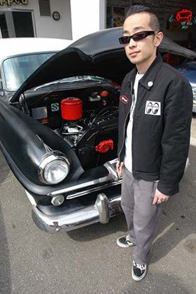 A man is standing in front of a black car with the hood up.