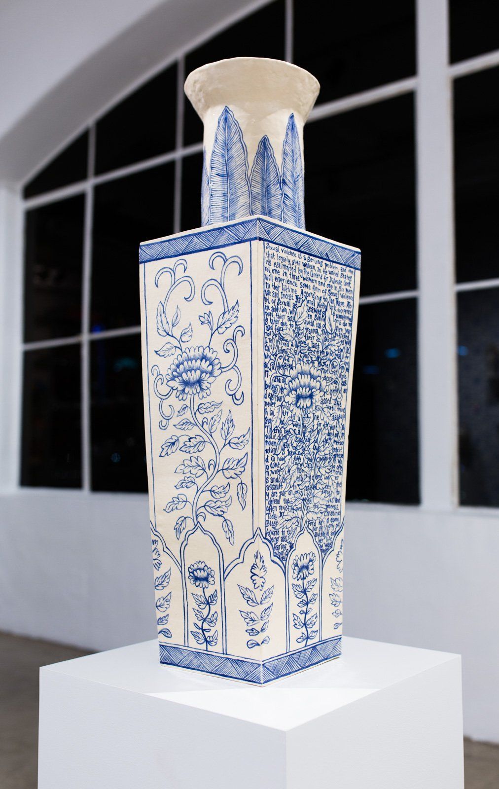 A blue and white vase is sitting on a white pedestal in front of a window.