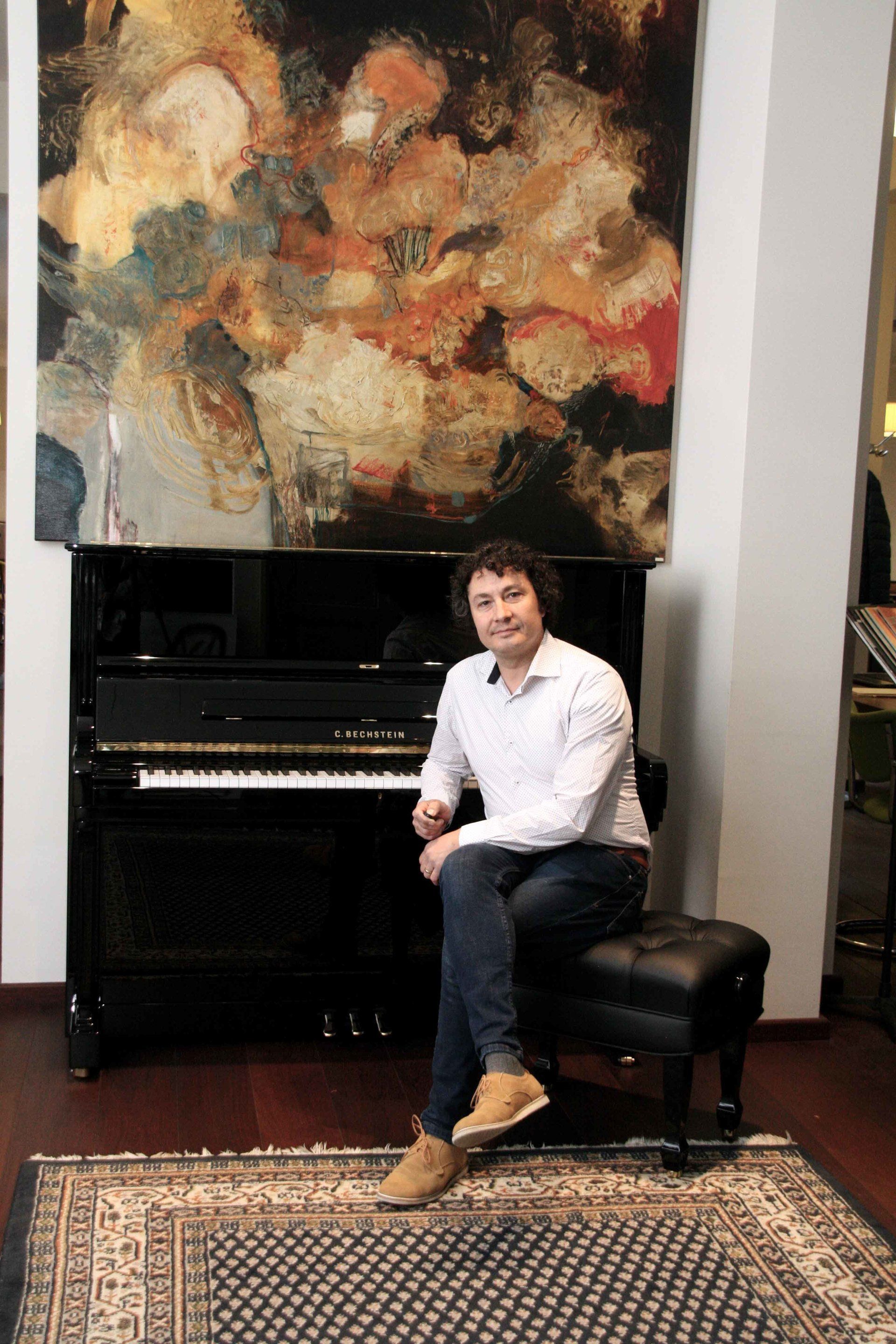 De pianotechnicus Popov zit op een pianokruk naast een C.Bechstein zwarte piano, onder een abstract schilderij.