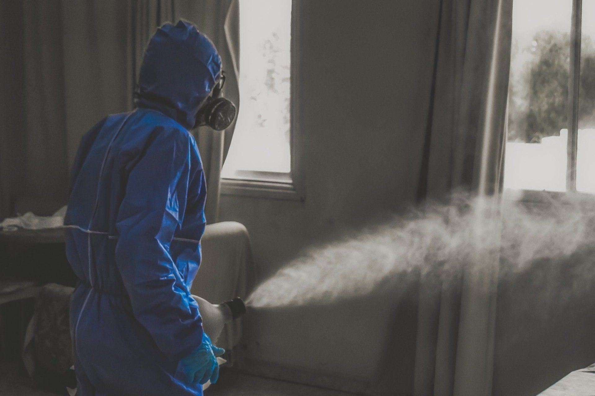 Person in blue hazmat suit sprays disinfectant in a room with a window.