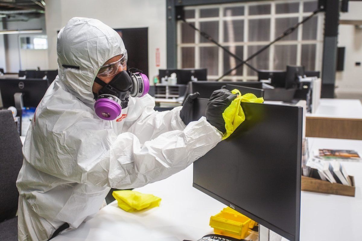 Person in hazmat suit disinfecting a computer monitor in an office setting.