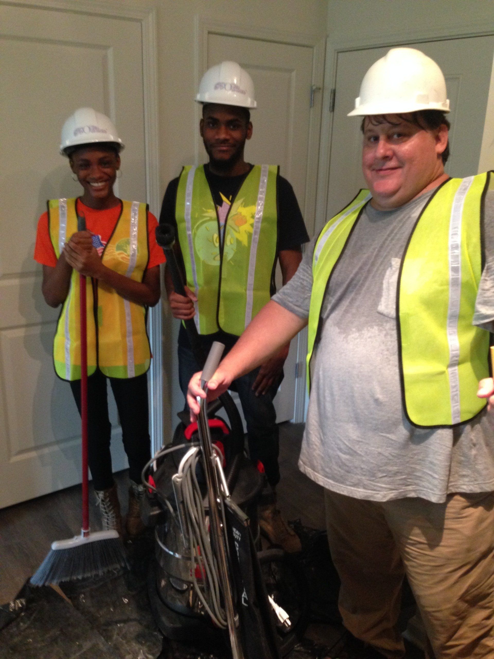 Three people wearing hard hats and safety vests cleaning up a room; two hold tools, one smiles.
