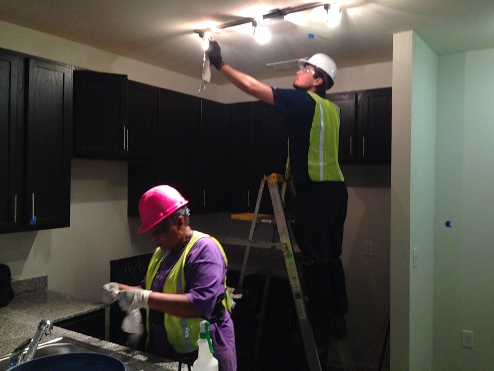 Two people in safety gear working in a kitchen: one on a ladder with lighting, the other cleaning the sink.