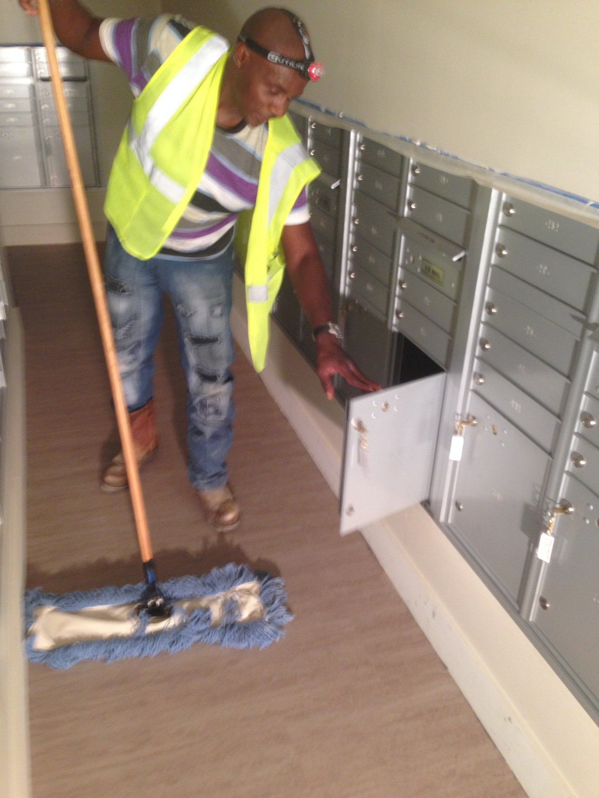 A person in a safety vest opens a mail slot with a mop beside them in a hallway.