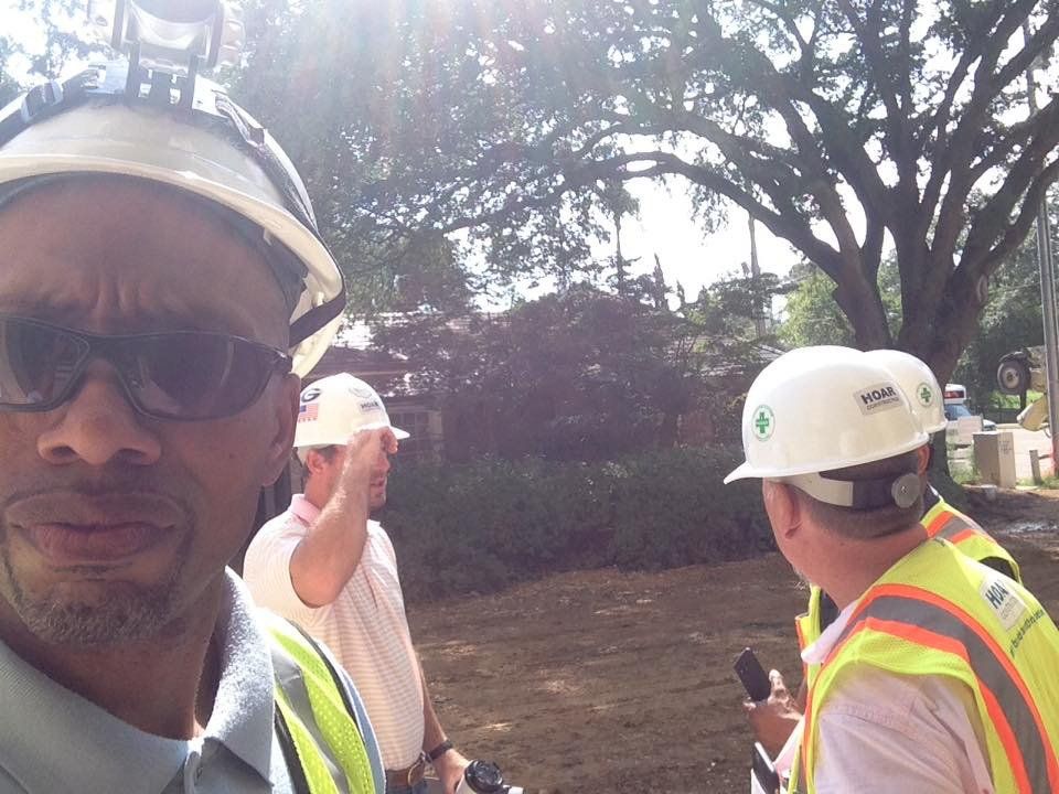 Men in safety vests and hard hats outdoors, one taking selfie. Sunny day, trees in background.