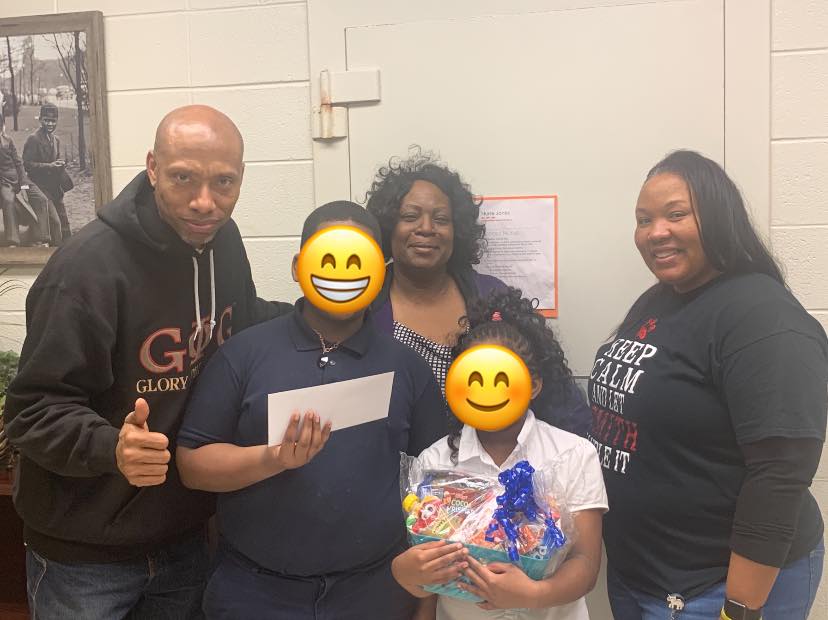 Group of five people indoors, two children, two adults, and a person holding a check. 