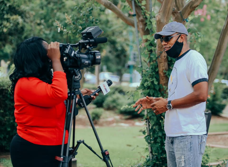 Woman filming man outdoors with video camera; both wearing masks.