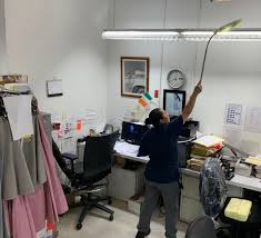 Person dusting a ceiling light in an office cluttered with paperwork, a desk, and hanging garments.