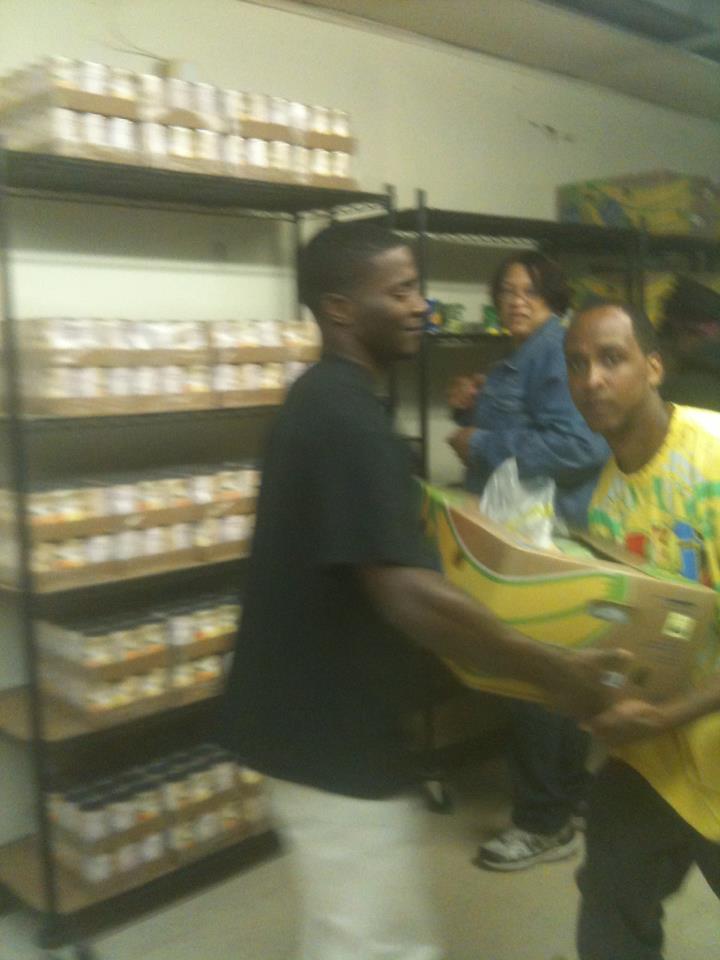 Two men and a woman in a room with shelves of items, one man carrying a box.
