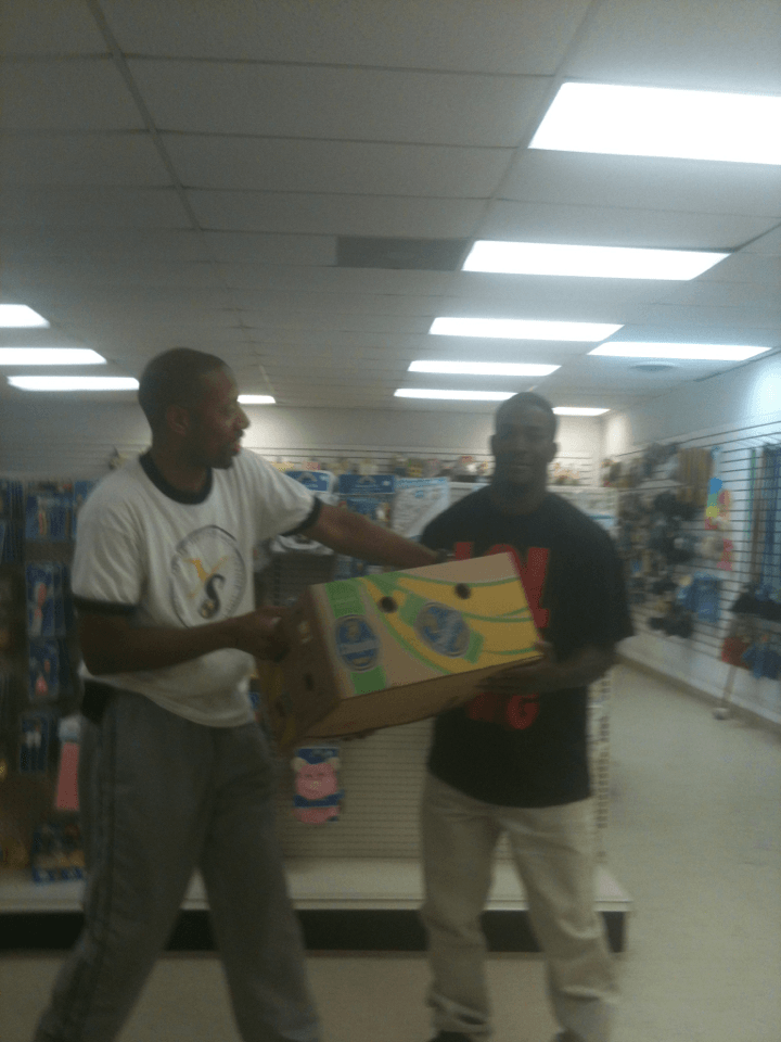 Two men holding a cardboard box in a store. One is pointing. Fluorescent lights are overhead.