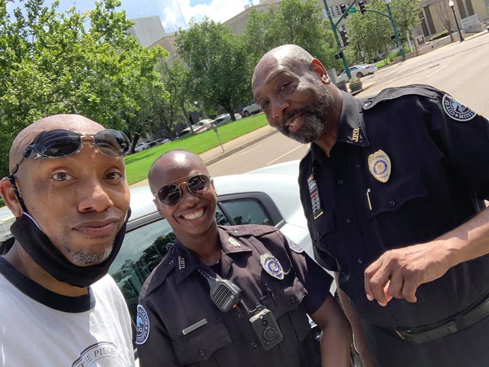 Three people posing for a photo outdoors. Two are in police uniforms, smiling. One holds the camera. 