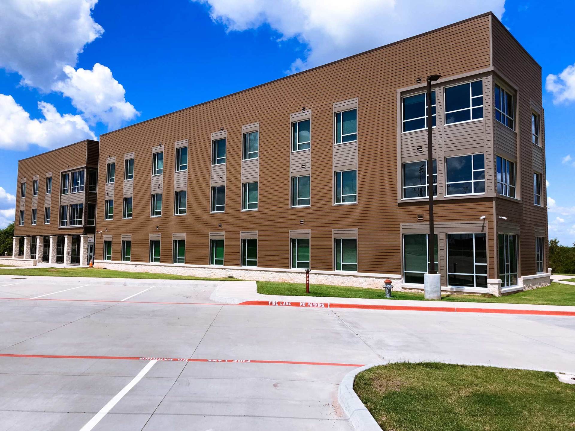 Modern three-story brick building with large windows beside an empty parking lot under a blue sky