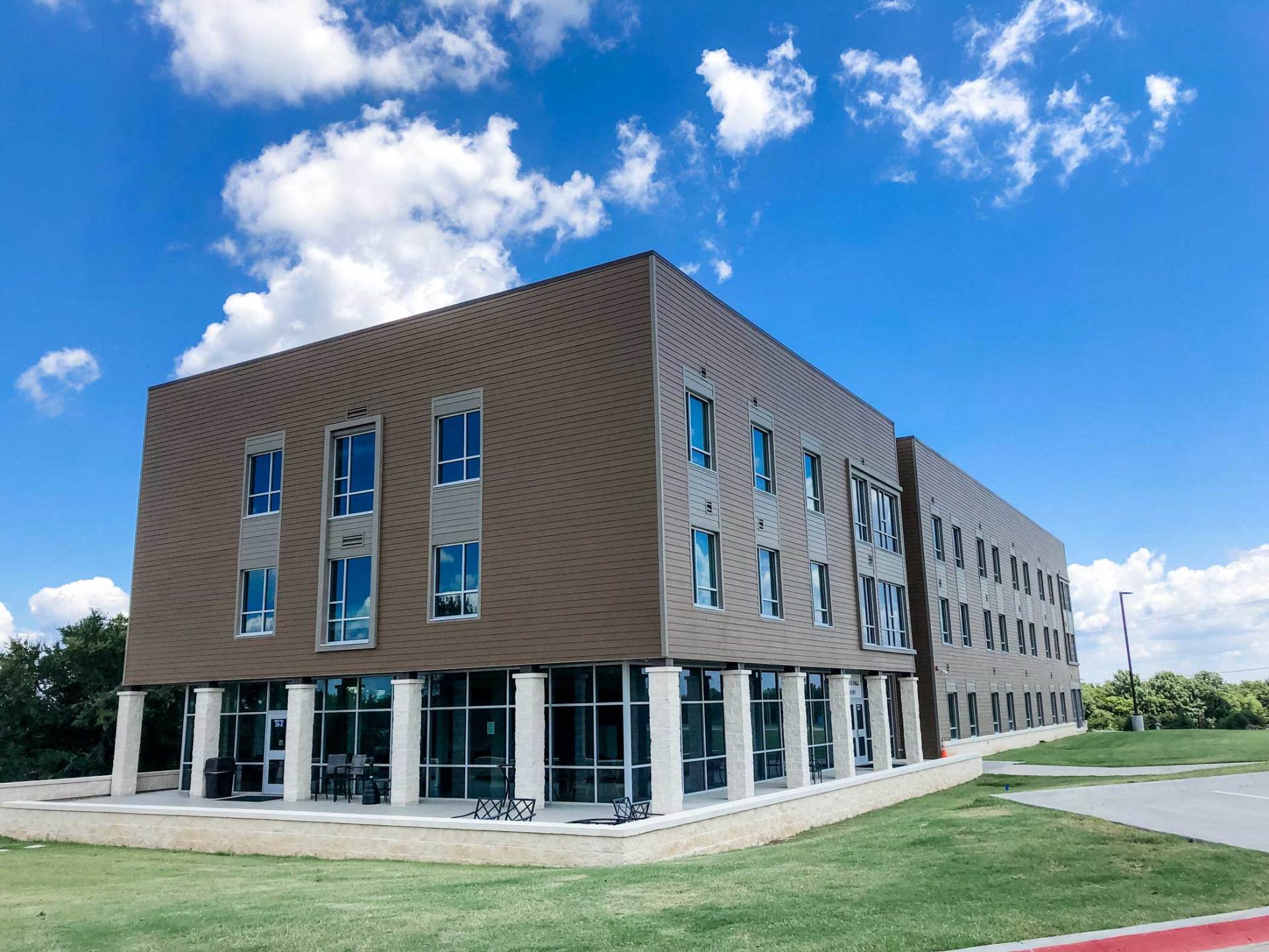 Modern three-story office building with brown siding, large windows, and a grassy lawn under a blue sky