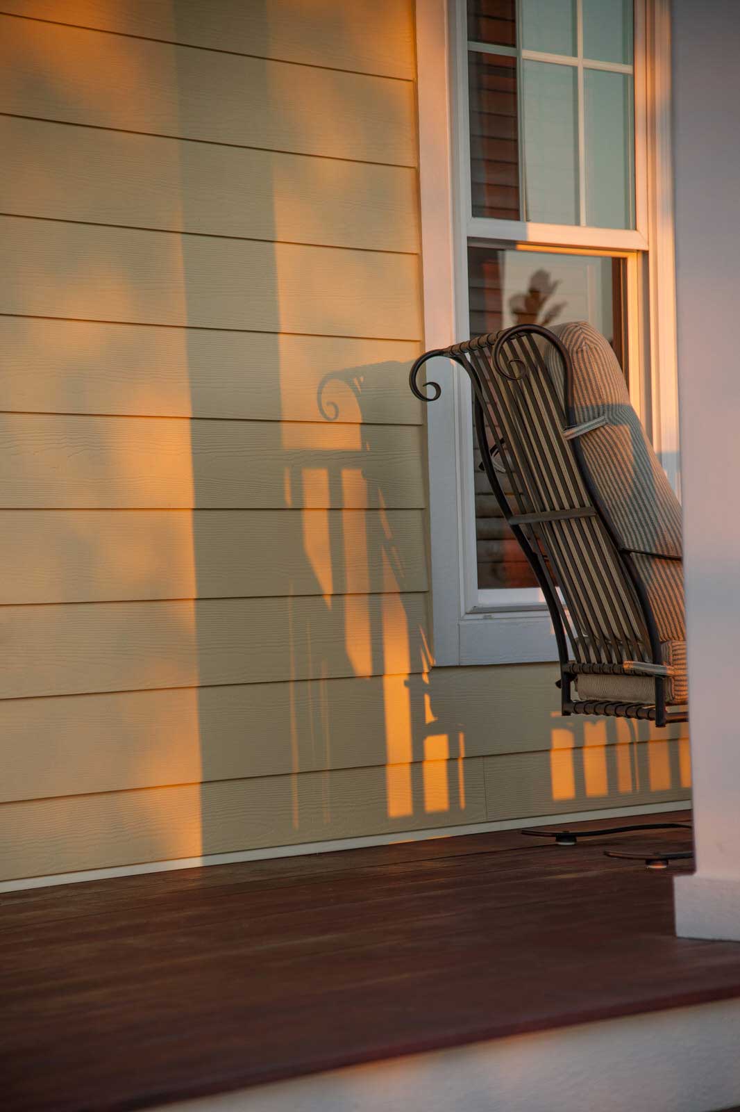 Wicker chair hanging on a sunlit porch beside a white-framed window and beige siding