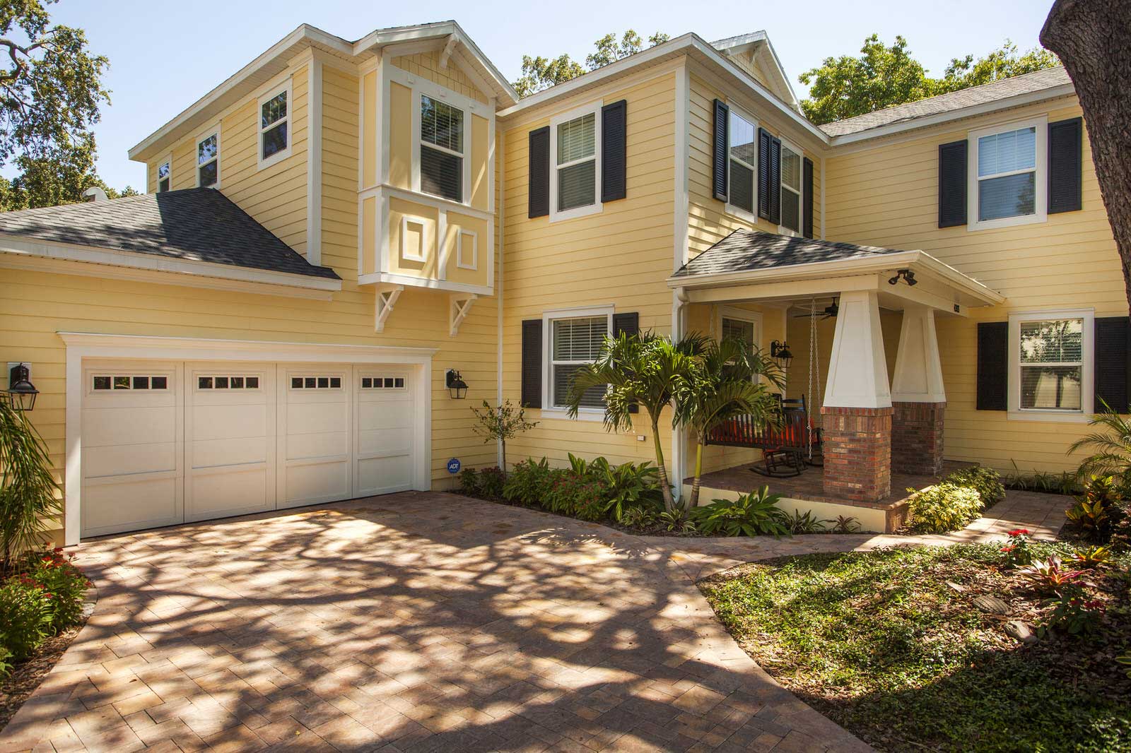 Yellow two-story suburban house with a two-car garage, front porch, and sunlit driveway surrounded by trees