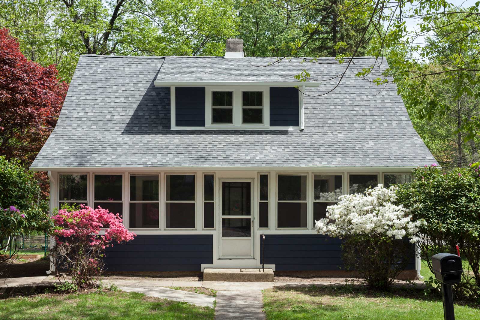 Small gray house with a screened porch, dark shutters, and flowering shrubs in front
