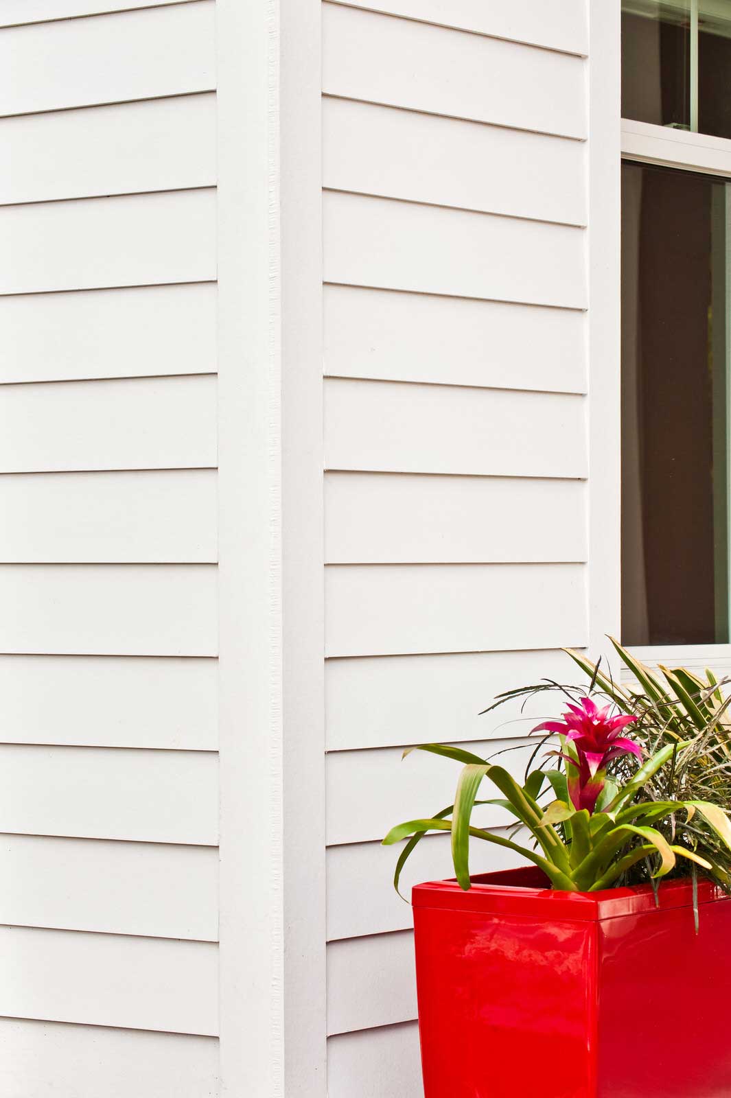 White house siding with a window and a bright red planter holding pink flowers and green leaves