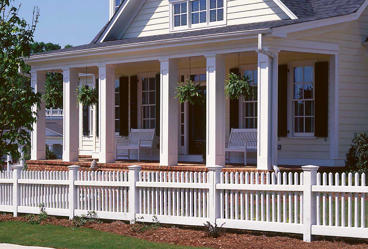 White house porch with columns, railings, hanging plants, and benches behind a white picket fence