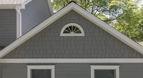 Gray house gable with small arched attic window and white trim under trees