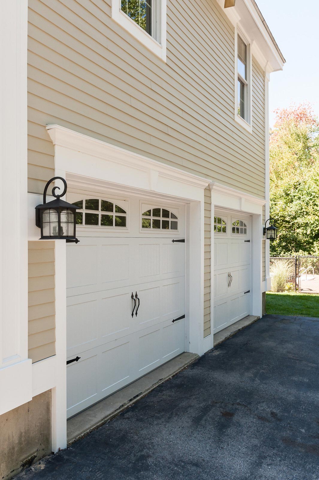 White garage doors on a beige house with a wall lantern beside a paved driveway.