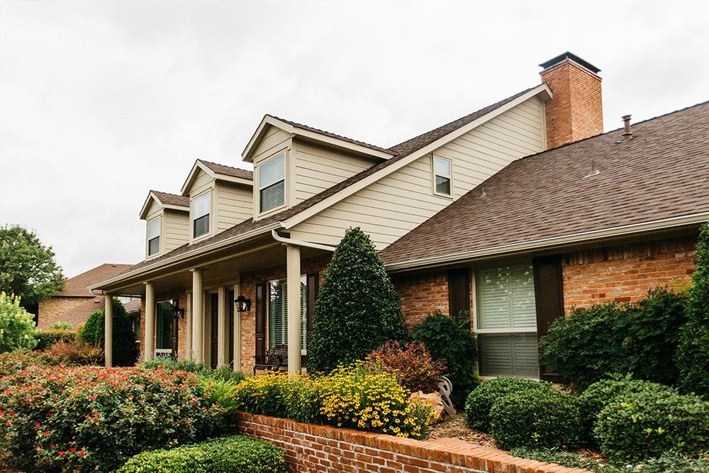 Suburban brick house with a covered porch, dormer windows, and landscaped shrubs and flowers