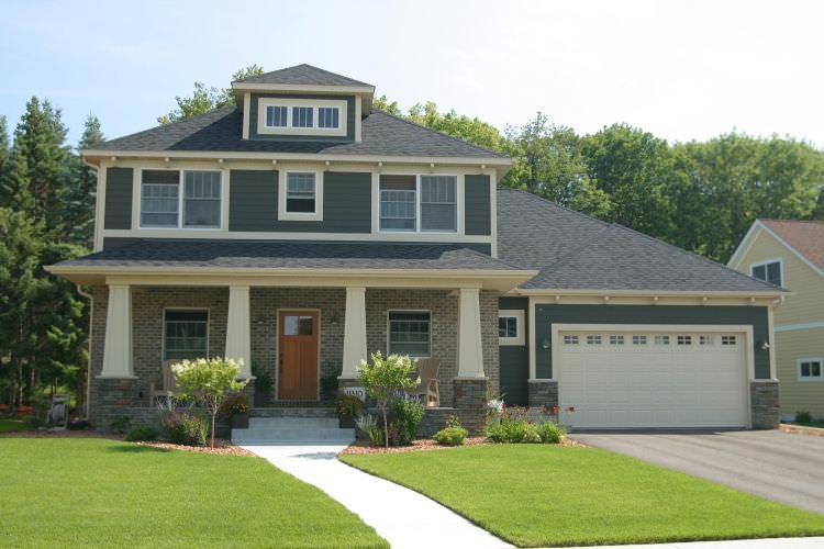 Two-story suburban house with a front porch, gray siding, and attached two-car garage, surrounded by a green lawn