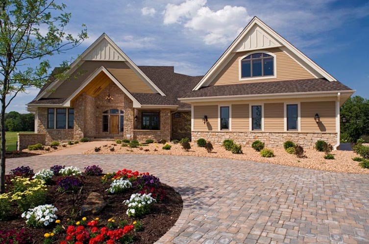 Large beige suburban house with stone entry, brick driveway, and landscaped front yard under a blue sky