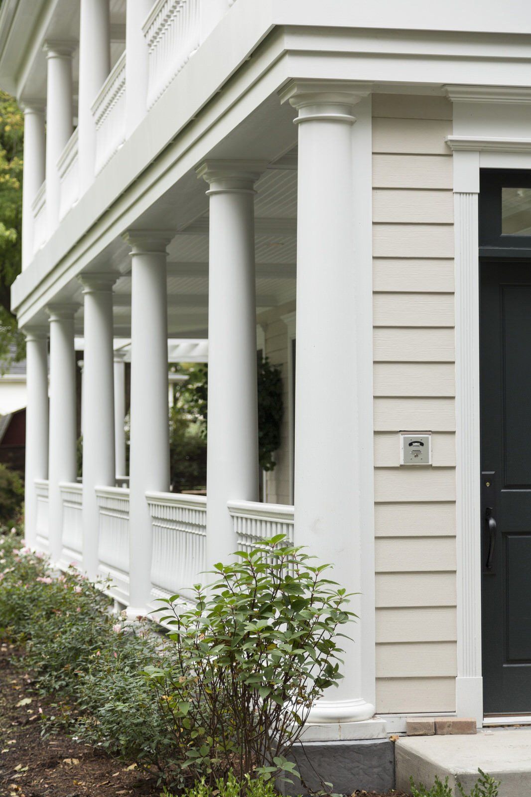 White-columned porch beside a dark front door and shrubs at a cream-colored house